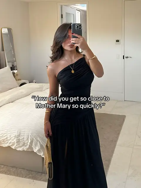 A woman is taking a mirror selfie in a bedroom. She's wearing a black, one-shoulder dress and holding a gold purse. The background includes a bed with white bedding, a mirror, and neutral-toned walls. Text overlayed on the image asks, "How did you get so close to Mother Mary so quickly?"