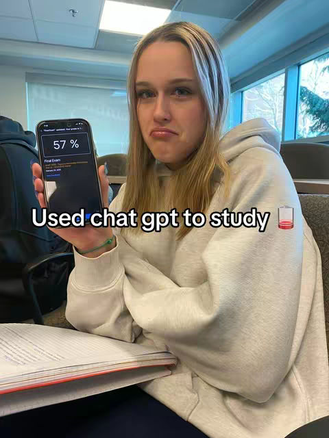 A young woman with long blonde hair sits in a classroom setting, holding a smartphone displaying a test score of 57%. She has a slightly distressed expression. In front of her is a stack of papers, likely textbooks or notes. The background includes rows of desks and windows. The image is well-lit, capturing the details of the woman and her surroundings.