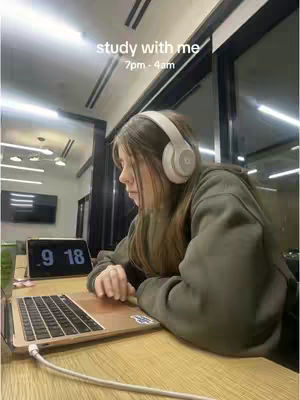 A young woman with long brown hair sits at a wooden desk, appearing to work on a laptop. She wears white headphones and a casual olive-green hoodie. An illuminated clock displays the time. The background includes a window and ceiling lights.