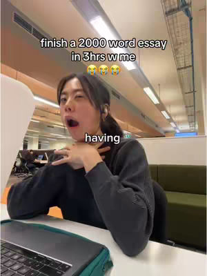 A young woman with dark hair is in a study environment with text overlayed on the image. She is mid-yawn with her hand covering her chest as if to stop the yawn. She has a laptop on a desk in front of her and the text says "finish a 2000 word essay in 3hrs w me", indicating a study vlog or tutorial.