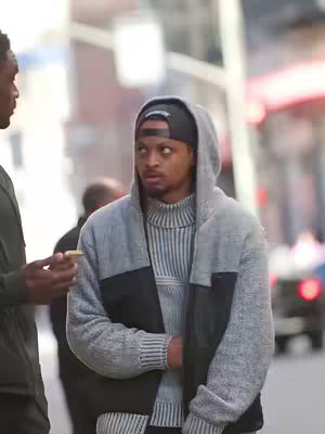 Two men are standing on a city street, one looking surprised. He is wearing a grey hoodie and a black beanie, with a perplexed look on his face. The other man is partially visible, with a watch on his left hand. The background is blurred, with hints of buildings and traffic.