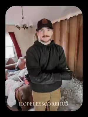 A man stands in a room that appears to be in disrepair. He is wearing a black hoodie, a baseball cap, and tan pants. Parts of the walls are visible with what look like curtains in the background, the ceiling is visible and there are many objects in the periphery of the room.