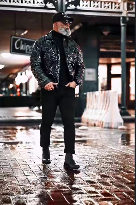 The image shows a man in a dark, urban setting. He is wearing a black, shiny jacket, a black turtleneck, black pants, and black boots. He is also wearing a black cap, sunglasses, and has a grey beard. He is standing on a brick-paved surface that appears wet, with reflective puddles. The background has blurred lights and a sign that says 'Chips'.