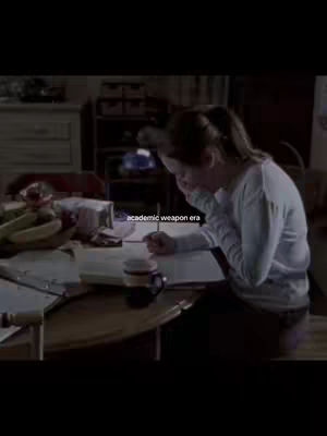 A woman sits at a table, illuminated by soft lighting, surrounded by books and a mug. She is writing, possibly studying in what seems to be cozy setting. The text overlayed says academic weapon era.