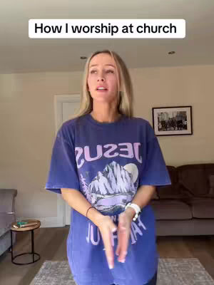 A woman stands in a living room setting, wearing a blue t-shirt that has the word 'JESUS' printed largely on the front, in an eye-catching purple color. The image includes a room with a window, a rug with a dark couch and side table in the background, and a framed picture on the wall. The woman is the main focus of the image.