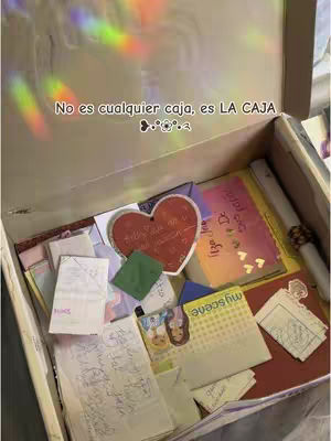 An open cardboard box filled with various items. Inside, viewers can see a large red heart-shaped card, smaller cards with colorful illustrations, a handwritten note, and a yellow notebook. The contents are layered, and the image is lit with soft, natural light.
