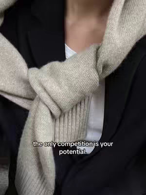 The image shows the upper body of a person, likely a woman, with a neutral background. She is wearing a white shirt underneath a black blazer. Wrapped around her neck is a tan sweater. Text overlayed on the image says, “the only competition is your potential.”