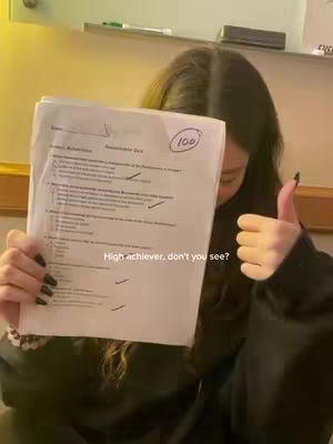 A young woman is holding up a test paper that has a perfect score of 100, with a thumbs-up gesture. The paper is held in front of her face, but the high score is clearly visible. The background includes a wall and a shelf.