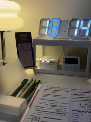A well-lit desk setup with a digital clock prominently displayed. The clock shows the time in blue LED digits. To the left, a lamp provides soft lighting. In the foreground, there's an open notebook with handwritten notes and pens, indicating study materials. A pen holder and other items also present, suggesting an organized workspace. The overall color scheme is neutral, with a focus on functionality and a clean environment.