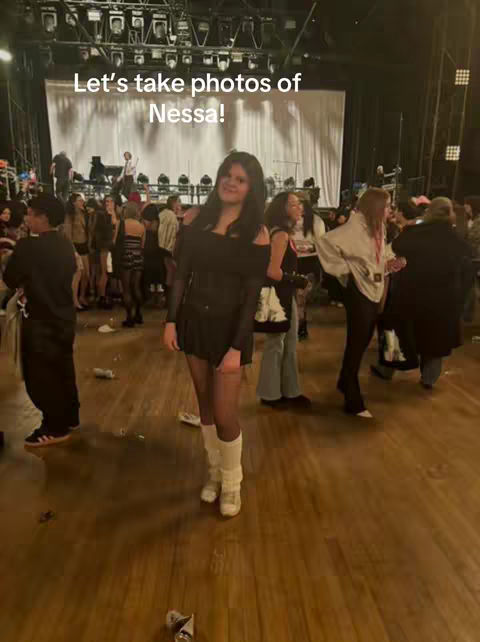 A candid shot taken in what appears to be a concert venue. In the foreground, a woman wearing a black outfit with off-the-shoulder sleeves, short skirt, and white boots gazes directly at the camera. Behind her, a crowd of people is visible, along with stage-like features, enhancing the backstage or pre-show atmosphere. The setting suggests a live event environment.