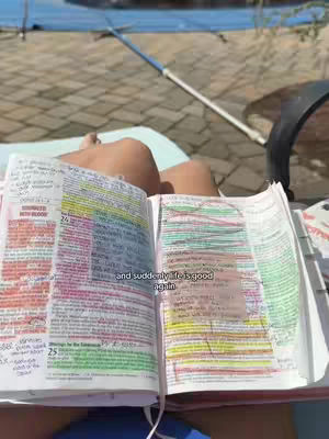An open Bible is shown, resting on what appears to be a tanning bed or lounge chair. The book is open, presenting an interior shot with highlighted text and handwritten notes. The background suggests a sunny outdoor setting, possibly near water.