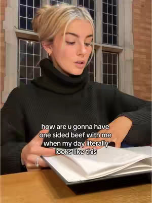 A young woman with blonde hair in a bun and a black turtleneck sits behind a desk, looking toward the camera while holding a book open in front of her. The background includes a window with multiple panes and a brick wall.