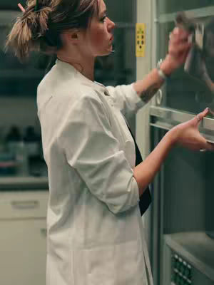 The image captures a woman in a lab setting, likely a scientist, wearing a white lab coat and with her hair up in a bun. She's holding a cloth and appears to be cleaning a glass panel, with a focused expression. The background shows a sterile lab environment with equipment.