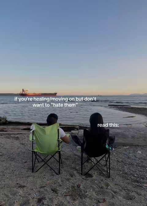 Two people, seen from behind, sit in folding chairs on a beach, facing the water. The sea stretches to the horizon, where a small ship can be seen. The sky is a gradient of blue and pink.