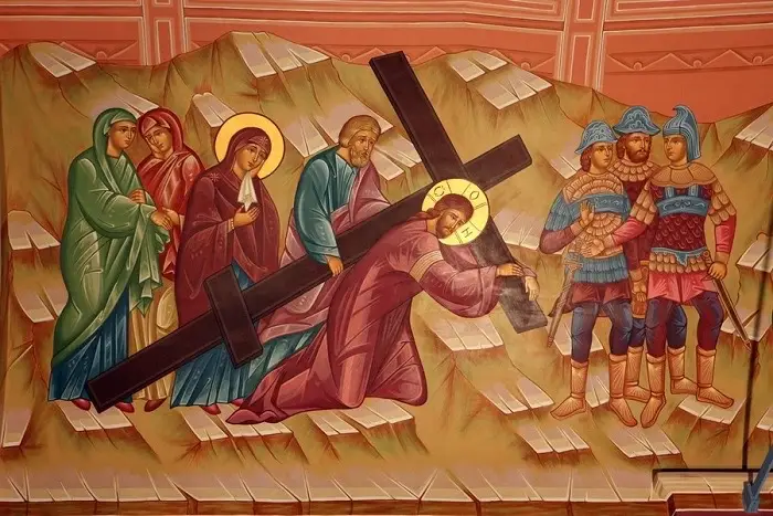 A religious painting depicting the crucifixion of Jesus Christ. Jesus is shown carrying the cross with other figures around him, including soldiers and women. The colors are muted, with a focus on blues, reds, and browns.