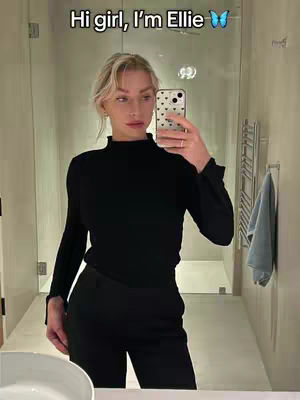A selfie of a woman in a well-lit bathroom. She is wearing a black mock turtleneck and black pants. She is holding a phone up to take the picture and has blonde hair. The background includes a mirror, a towel rack, and a sink.