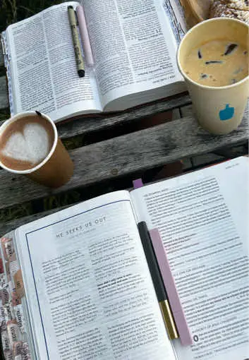 The image shows a close-up shot of items on a wooden surface, likely a table. There are three open books with text and pens. A cup of coffee with a heart-shaped design in the foam is placed near one of the books. Two additional cups with beverages are visible, one with what appears to be coffee and the other with a lighter drink. All are placed on a rustic wooden surface, and the overall composition suggests a setting for reading, study, or reflection.