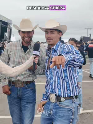 Two men in cowboy hats and casual attire are standing in an outdoor setting. One holds a microphone and the other points at the camera. Text overlayed in a pink shape says 'DESCARGA CHISPA' and appears to be a call to action for an app.