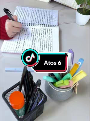 The cover image shows a top-down view of a desk with a notebook open, a hand writing, and various stationery items. The background is a light surface, possibly a desk or counter. In the foreground, there's a notebook open to a page with handwritten text. A hand holding a pen is visible, actively writing on the notebook. To the right, there are several colorful highlighters in a small gray container. A black pen, a small container with an orange label, and other smaller stationery items are also visible, arranged in a black mesh container. The center of the image includes a TikTok overlay with the text "Atos 6". The color palette is warm, with a mix of bright colors from the highlighters and the red sleeve of the person writing.