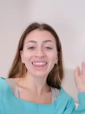 A smiling woman with brown hair and blue clothing sticks her tongue out, posing in front of a plain backdrop. She's wearing small gold hoop earrings and has a light skin tone, with natural makeup.
