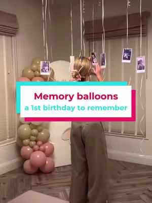 A woman stands in a room, facing away from the camera in front of a large balloon display. Balloons in shades of gold and pink adorn the background. Hanging from above are photos, each suspended by a string. Light streams in from a window on the right. The foreground shows a portion of the floor and a cabinet.