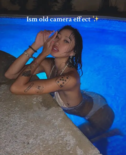A woman is leaning against the edge of a pool, with her body submerged in the illuminated blue water. She has tattoos on her arms and is wearing a bikini top. Text overlay indicates an old camera effect.
