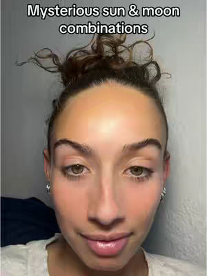 A selfie-style shot featuring a person with their hair pulled up in a bun and visible makeup. The individual is looking at the camera with a slight smile. In the background, a plain wall is visible.