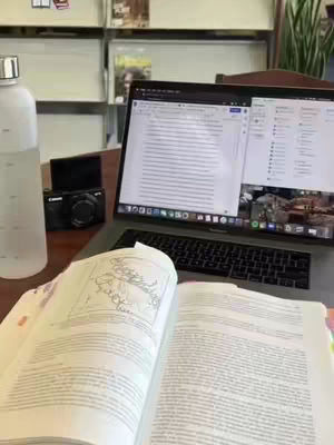 An open book is in the foreground, with the pages facing up. A laptop is in the background with its screen open and facing towards the camera. A water bottle and a camera are on the surface in front of what appears to be a bookshelf.