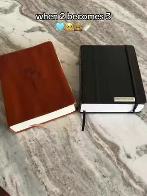 The image showcases two closed books, one reddish-brown with a heart-shaped design and the other black. They are positioned on a marble-patterned surface with the text overlayed at the top: 'when 2 becomes 3' accompanied by a blue heart, surprised face, teddy bear and baby bottle.