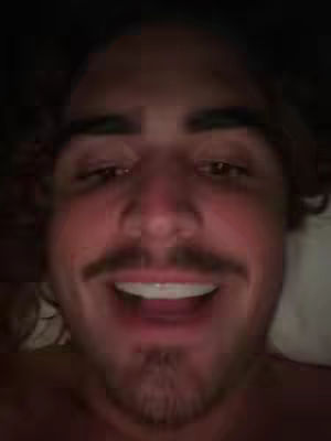A selfie-style shot of a man's face in low lighting. He has dark curly hair, a mustache, and a goatee. He is smiling widely with his mouth open. The background is dark.