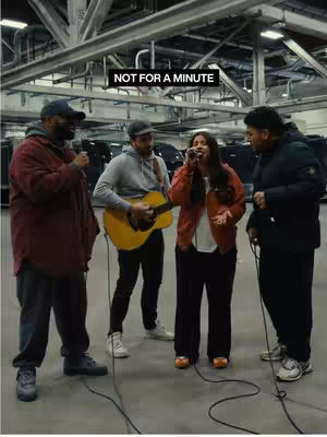 Four people in a large hall. One woman sings with a microphone in her hand while another man plays the guitar. The other two men are standing with a microphone in their hand, watching. The background is a large open space with metal structures.