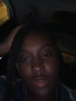 A low-angle selfie shows a woman in a car, filling the frame with her face. Her hair is dark and parted on the side. She is looking directly at the camera, with soft lighting casting shadows. The background suggests a dimly lit interior, possibly a car or a room.