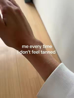 The video shows a person's lower arm and hand, with a white-sleeved wrist on display. The arm has a tanned appearance, and is positioned over a light-colored wooden surface. Text overlayed says 'me every time, I don't feel tanned'. The composition focuses on the arm, suggesting a desire for a tanned look.