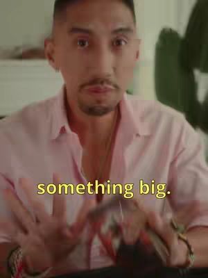 A man in a pink button-down shirt is holding and looking at a deck of tarot cards. The shot is from the mid-chest up. A yellow text overlay reads 'something big'.