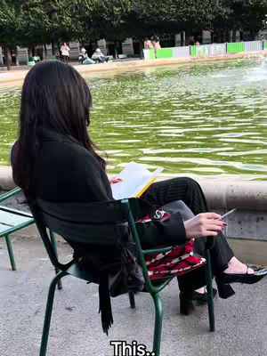 A woman sits in a green chair by a body of water. She is facing away from the camera, wearing a black outfit and holding a book and cigarette. In the background, there are trees, other people, and a green and white fence.