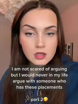 A close-up shot of a young woman with brown hair. She has fair skin, prominent eyebrows, and her lips are slightly parted. The background is blurred, but hints of an indoor setting with a light fixture are visible.