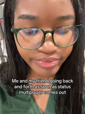 A close-up shot features a person with dark skin wearing green-framed glasses. The person's gaze is directed downward towards the bottom of the frame. Text overlayed on the image reads, 'Me and my friend going back and forth as soon as status multiplayer comes out.' The background appears blurred, with indications of an indoor setting.