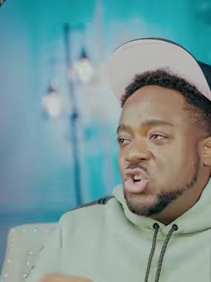The cover image shows a man in a light green hoodie and a white and black baseball cap, mid-sentence with a serious expression. The background is blurred, with hints of blue and white lights.