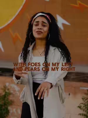 A woman with long, dark curly hair stands in front of a wall with colorful abstract art. She wears a white top, a light-colored coat or jacket, black pants, and a white headband. Text overlayed on the image says "WITH FOES ON MY LEFT AND PEARS ON MY RIGHT".