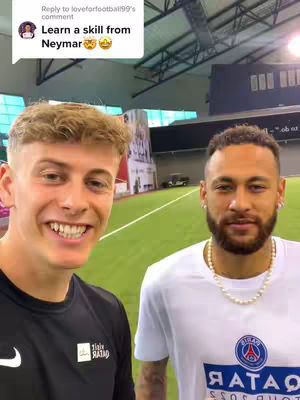 Two men are standing in a room; one is facing the camera and smiling, while the other wears a white t-shirt and stands slightly behind him. The room has a green turf floor and a tall background.