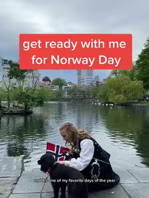 A woman and her dog are by a lake with buildings in the background. She is holding a Norwegian flag.