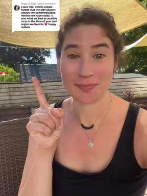A woman with light skin and brown curly hair is in an outdoor setting, pointing upwards with her right index finger and smiling. There's a backdrop of foliage and a partial view of a canopy above. She's wearing a black tank top and a necklace with a pendant. A text bubble is visible on the upper left of the frame.