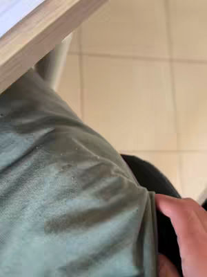 The cover image is a low-angle shot, offering a close-up view of a person's body from the waist down. A green garment, possibly a shirt or jacket, is draped over the body, with a hand visible at the center right. The muted green color of the clothing contrasts with the lighter tan tones of a surface, possibly the floor, and the wooden edge of a table in the upper left corner. This placement of elements suggests an interior setting with basic functionality.
