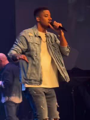 A man with a microphone in a performance setting, wearing a denim jacket over a white t-shirt and distressed jeans. He's gesturing with his hands, and another person is partially visible in the background, possibly a stage setup with dim lighting.