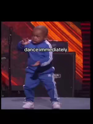 A baby wearing a blue Adidas tracksuit is dancing on stage. The baby is facing forward with arms out, as though performing. The background has a red backdrop.