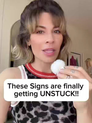 A woman with light brown hair is centered in the frame, facing the camera and speaking. She holds a white, fluffy object in her right hand. A text overlay at the bottom reads, "These Signs are finally getting UNSTUCK!!" The background out of focus with a painting.