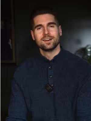 A man in a blue shirt looks towards the camera, with a dark background. The frame is a medium shot focusing on his upper body. There is a microphone on his shirt. A framed picture is visible in the background.