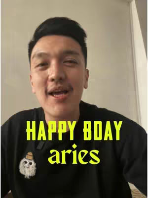 A man with dark hair is centered in the frame, with a blurred background suggesting an indoor setting. He wears a black shirt. The overlay text in a bold, yellow font reads \"HAPPY BDAY Aries\"