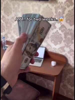 A person's hand holding up a large wad of US hundred-dollar bills, with the text "APT.. for two weeks.." overlaid on the image. In the background is a brown desk with a laptop and a small white object on top, and a patterned wallpaper.