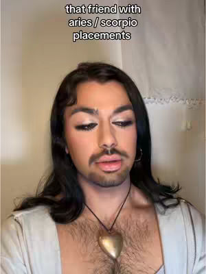 A close-up shot of a person with long dark hair and makeup, looking at the camera. The person has a mustache and a beaded necklace.